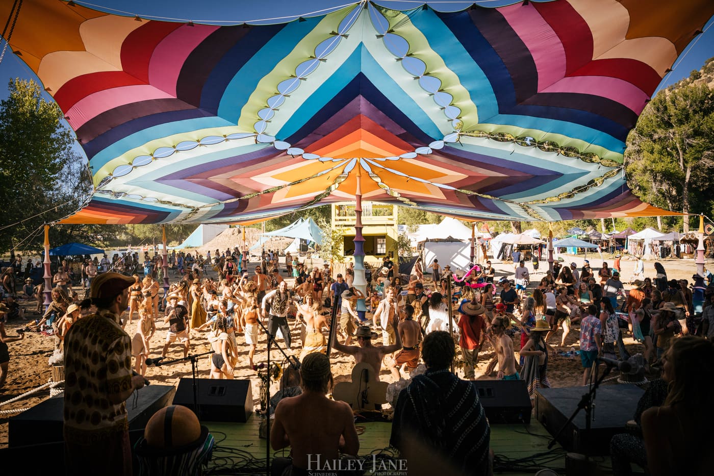 SANJI on the main stage at Unison Festival under a rainbow shade canopy, seen from behind the band, with a dancing crowd in the sun.